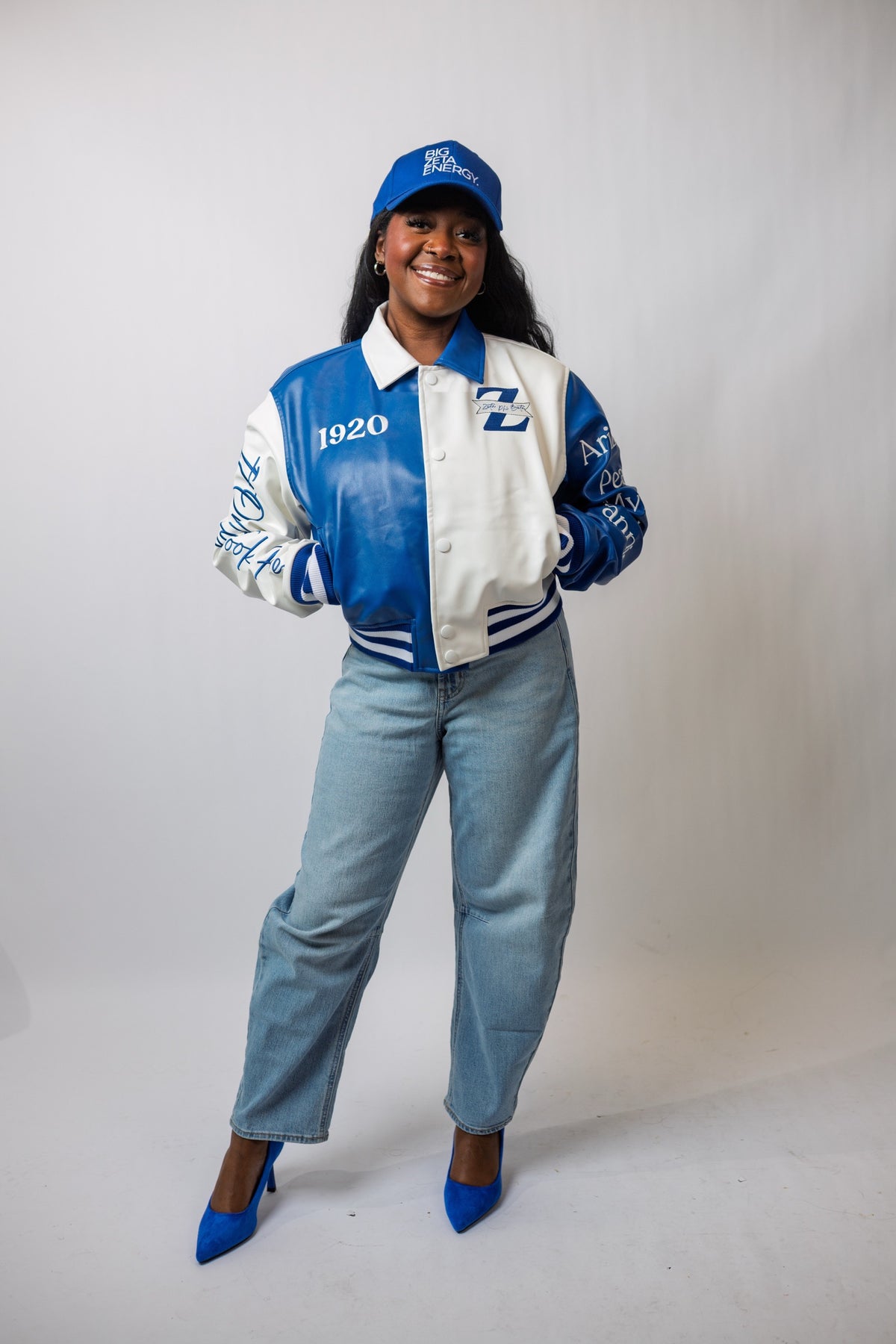 Model in the 5 For the Founders Leather Jacket, full length, with a Zeta cap, light wash jeans and Royal Blue heels