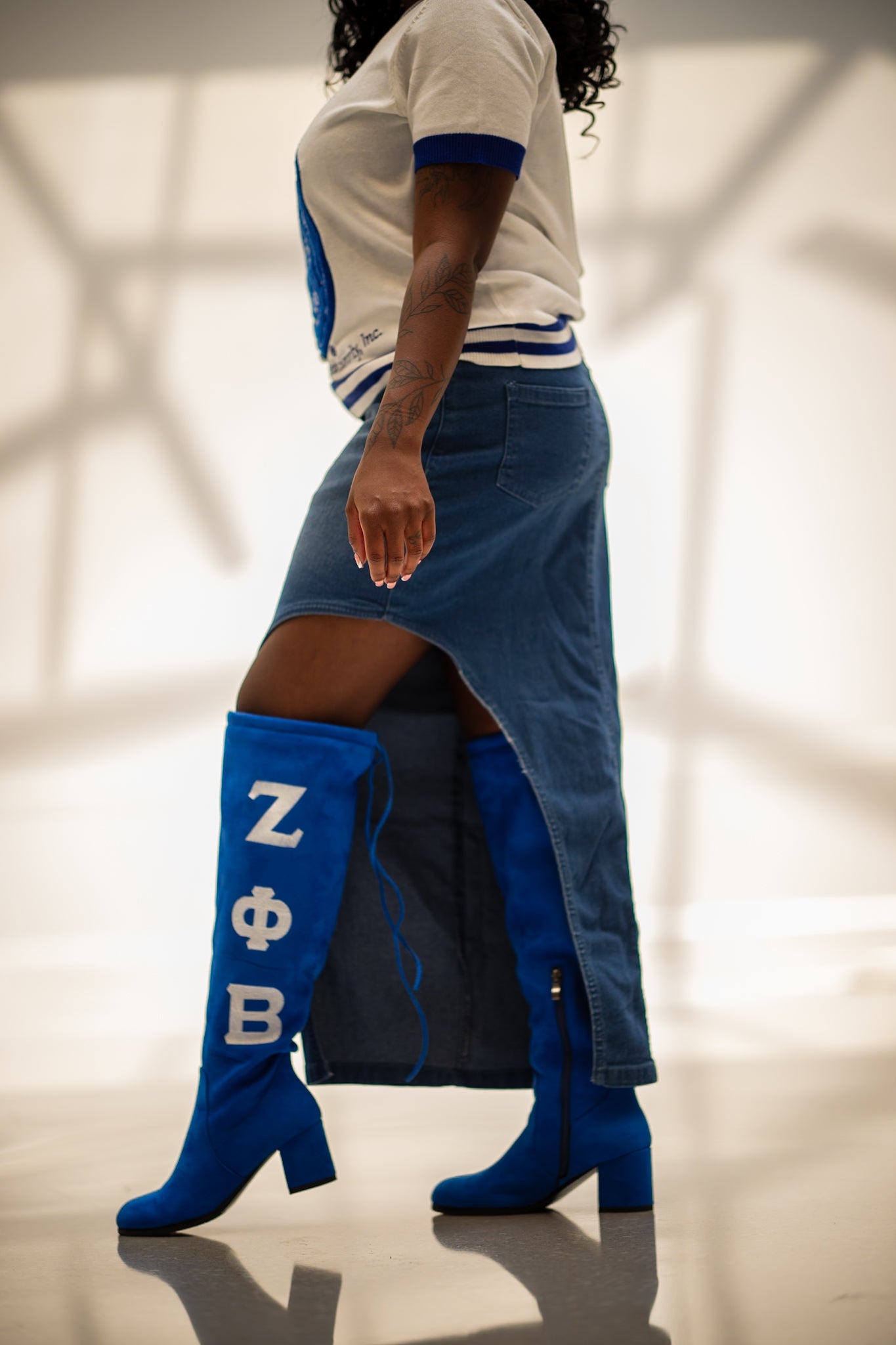 A model is shown from the side wearing a white and blue ringer t-shirt, a blue denim skirt, and bright blue heeled boots with white Greek letters 'Z Phi Beta' printed on the side.