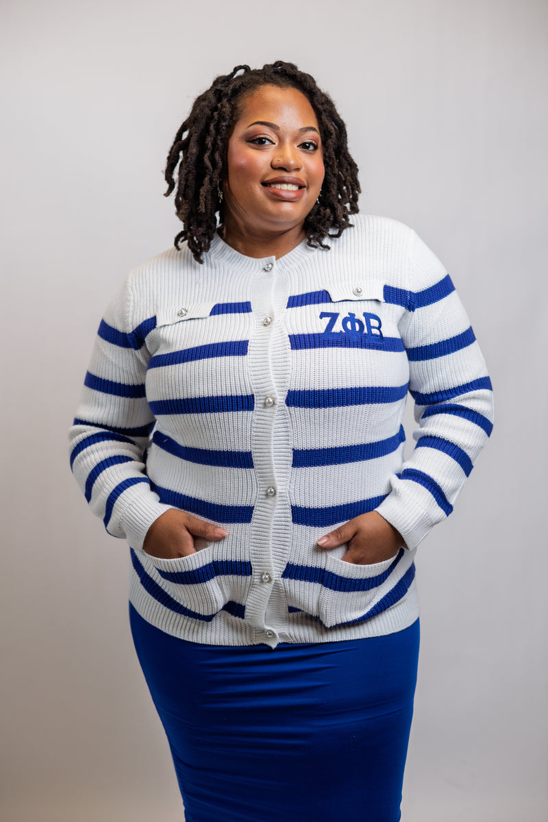 A woman wearing a white and blue horizontally striped cardigan with the Greek letters 'ZΦB' on the chest, paired with a solid blue skirt, standing against a light gray studio background.