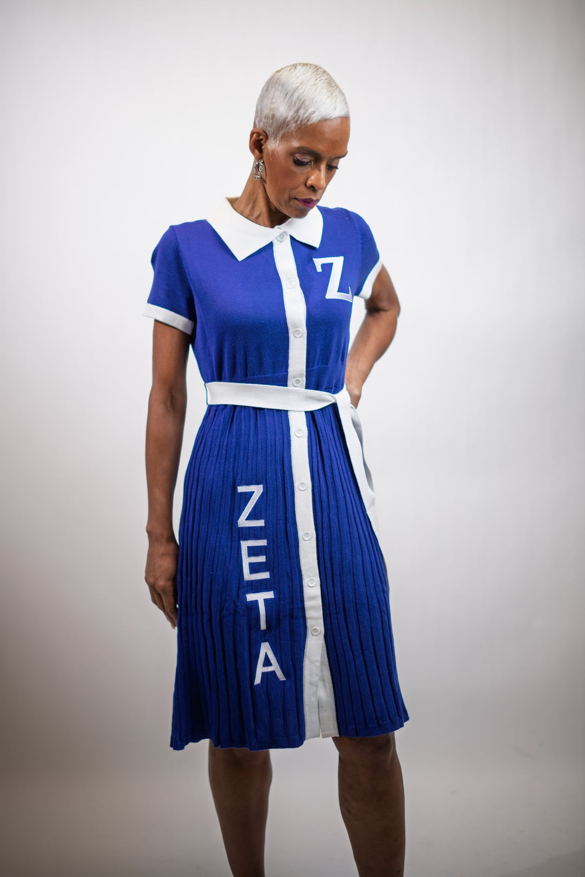 A woman with short silver hair wearing a royal blue and white pleated dress with the Greek letter 'Z' on the chest and 'ZETA' printed vertically on the skirt, posed against a neutral light gray background.