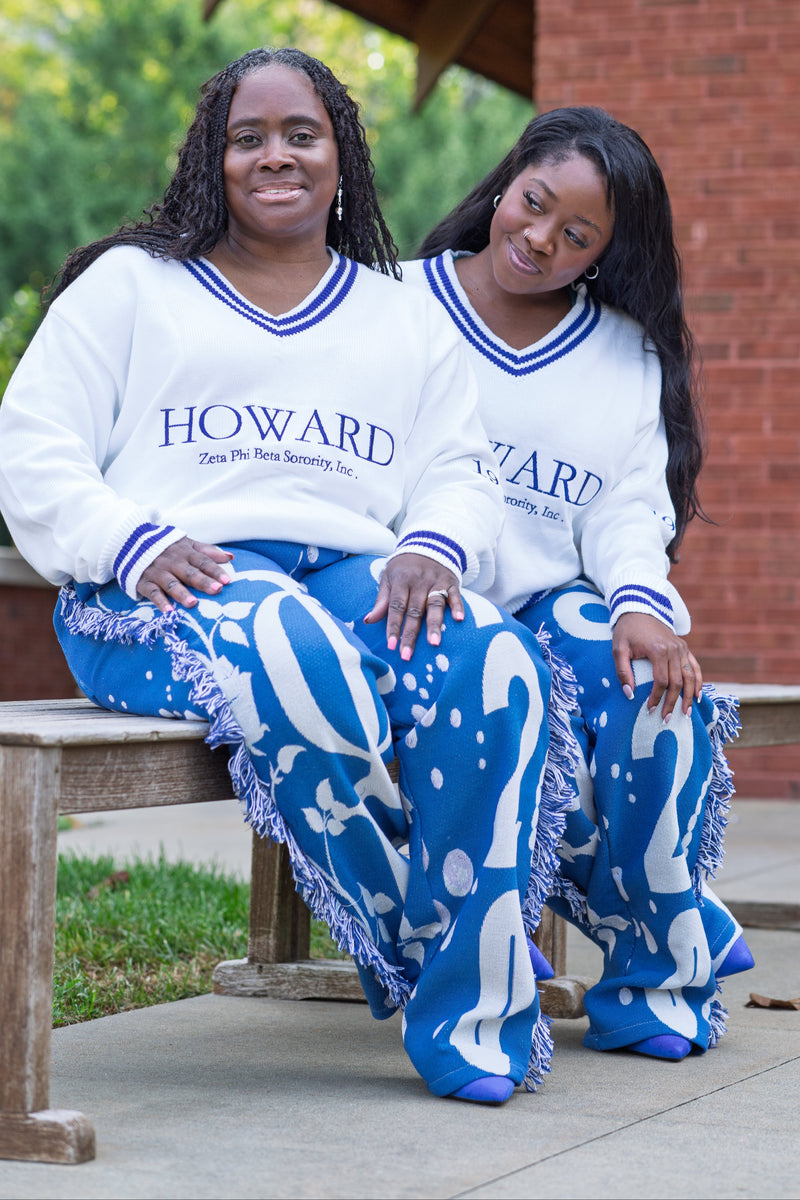 Two women sitting on a wooden bench outdoors, wearing matching white V-neck sweatshirts with blue trim and the word 'HOWARD' across the chest, paired with blue and white patterned fringed pants.