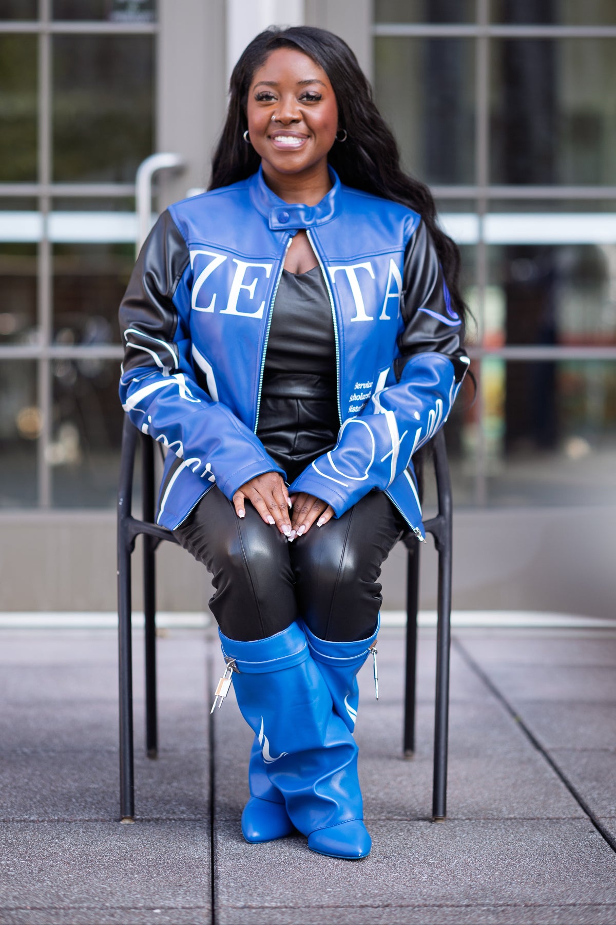 A woman sitting on a black metal chair outdoors in front of a modern glass building. She is wearing a blue and black varsity-style jacket with 'ZETA' lettering, a black top, black pants, and bright blue knee-high boots.