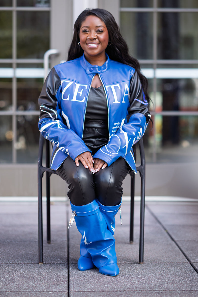 A woman sitting on a black metal chair outdoors in front of a modern glass building. She is wearing a blue and black varsity-style jacket with 'ZETA' lettering, a black top, black pants, and bright blue knee-high boots.