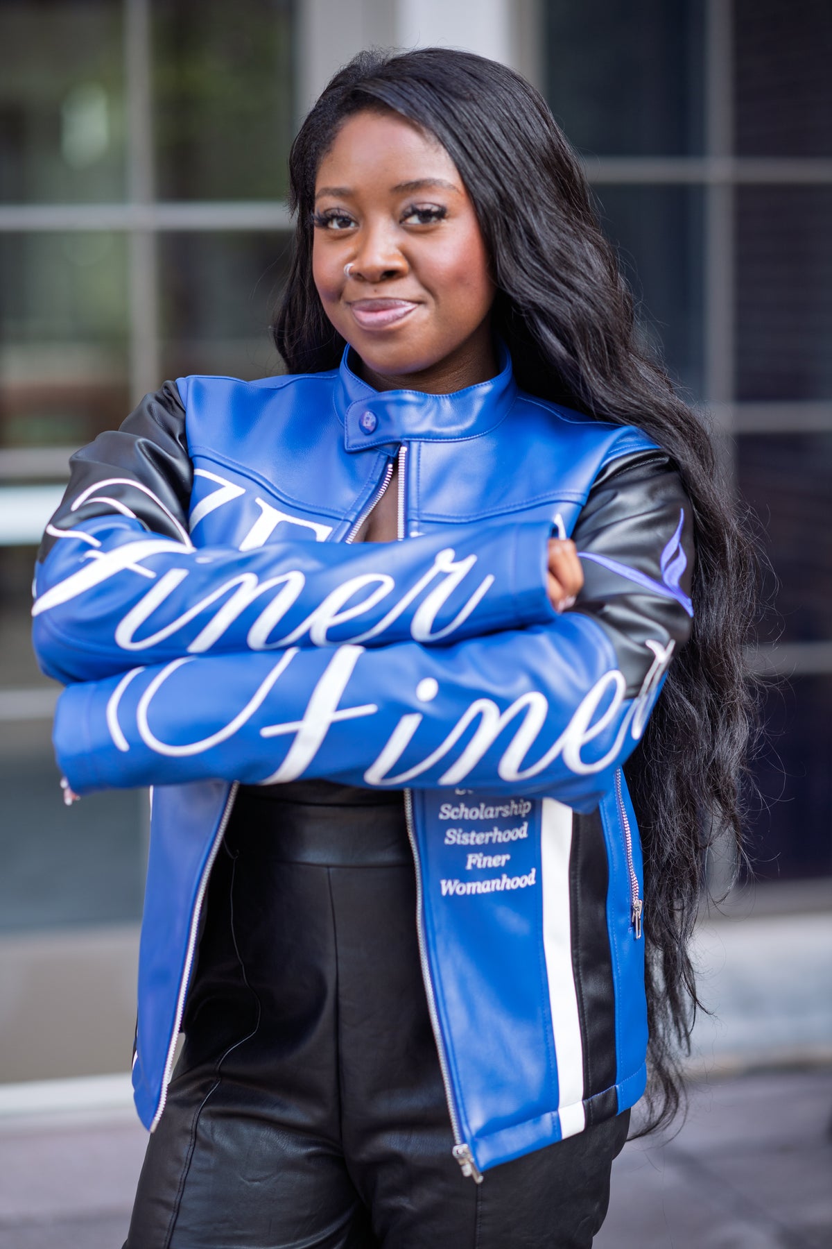 A woman with long black hair stands outdoors with her arms crossed, wearing a blue and black faux-leather letterman-style jacket with white script text reading 'Finer Finer' and black leather pants.