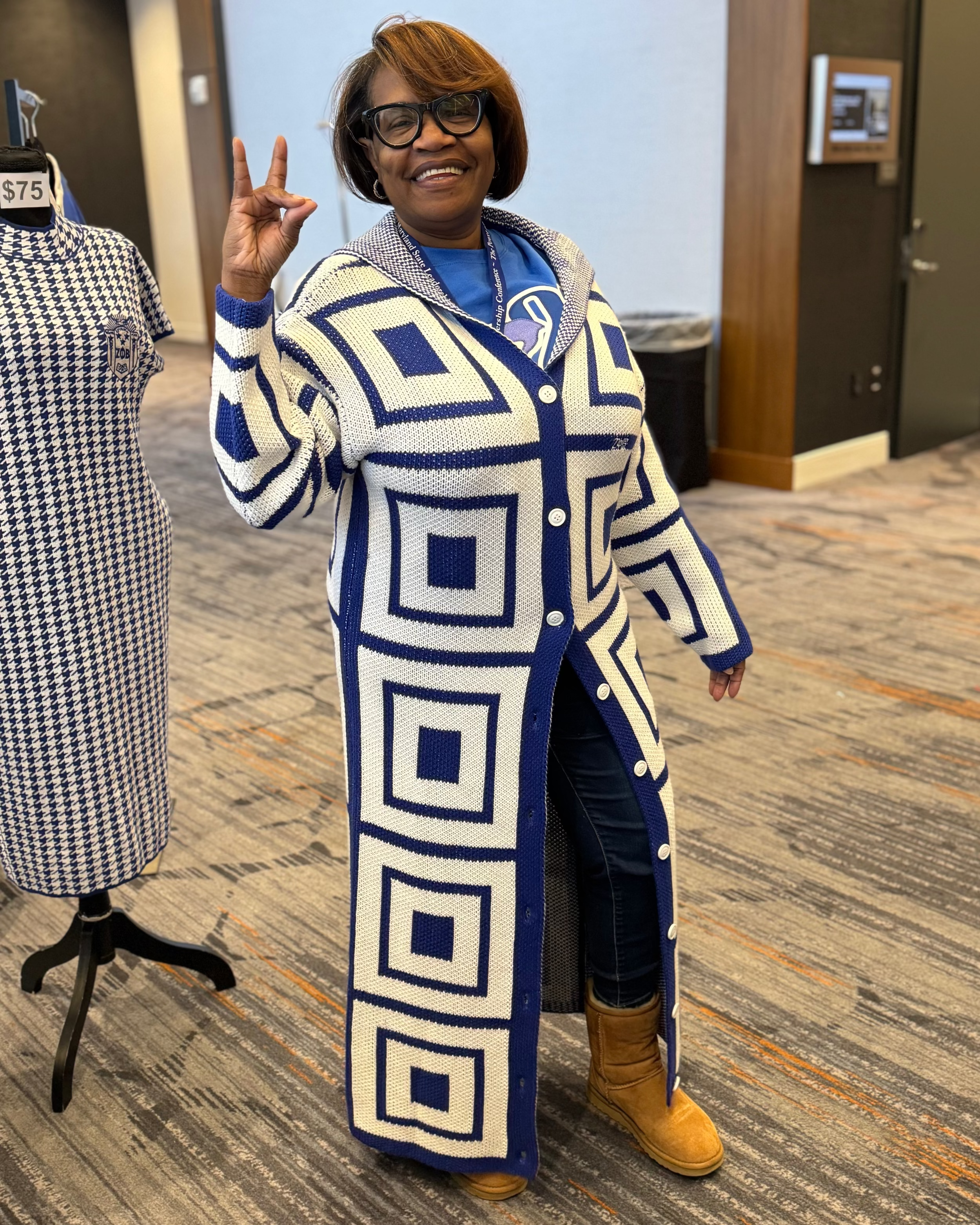 Woman wearing a long, Zeta Phi Beta patterned cardigan in an indoor setting with a mannequin and clothing display in the background.