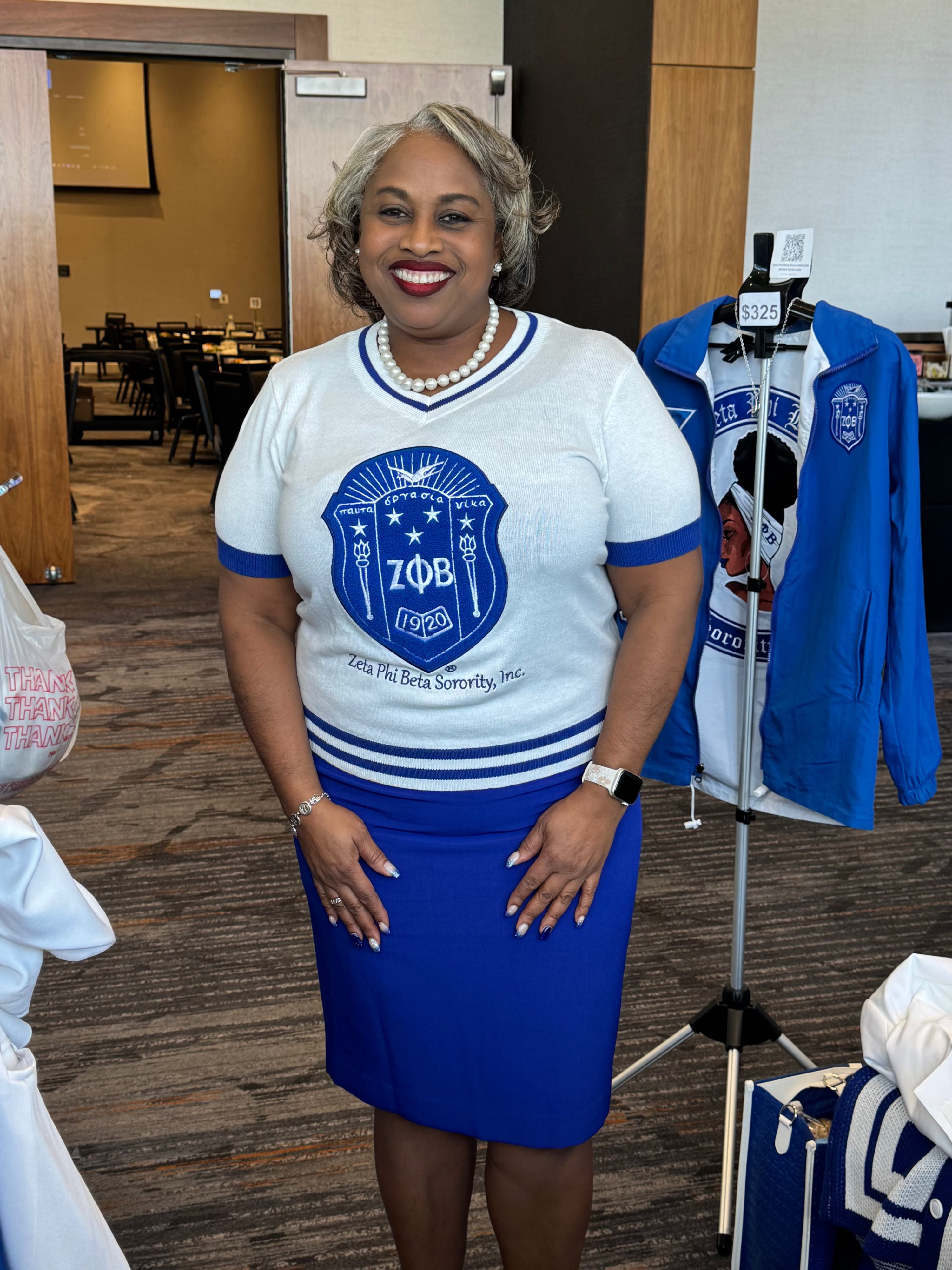 Woman wearing a white and blue Zeta Phi Beta sweater shirt, standing in an indoor setting.